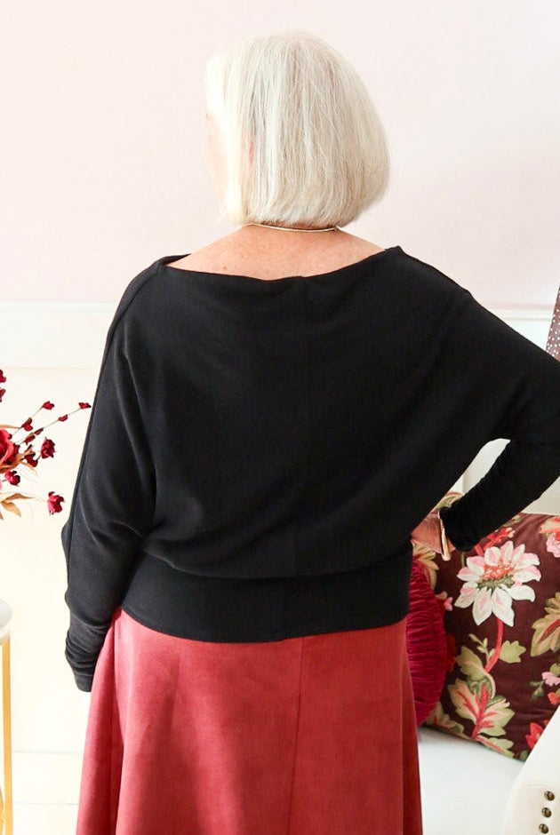 Woman wearing a black Kira knit top with boat neckline and relaxed dolman sleeves, styled with a burgundy suede skirt.