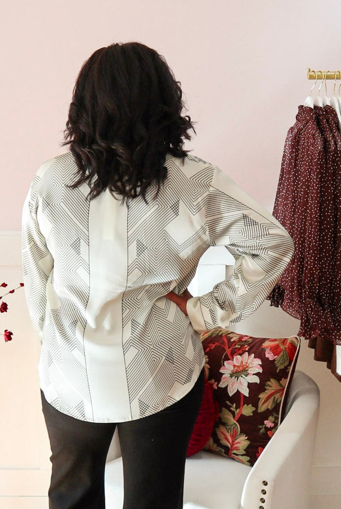 Woman wearing a cream blouse with black geometric print, classic collar, concealed button front, and long sleeves, styled with black denim jeans.
