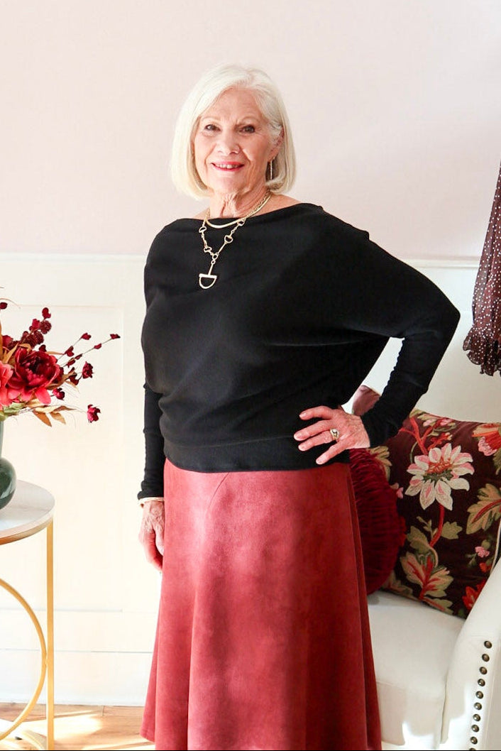Woman wearing a black Kira knit top with boat neckline and relaxed dolman sleeves, styled with a burgundy suede A-line skirt.