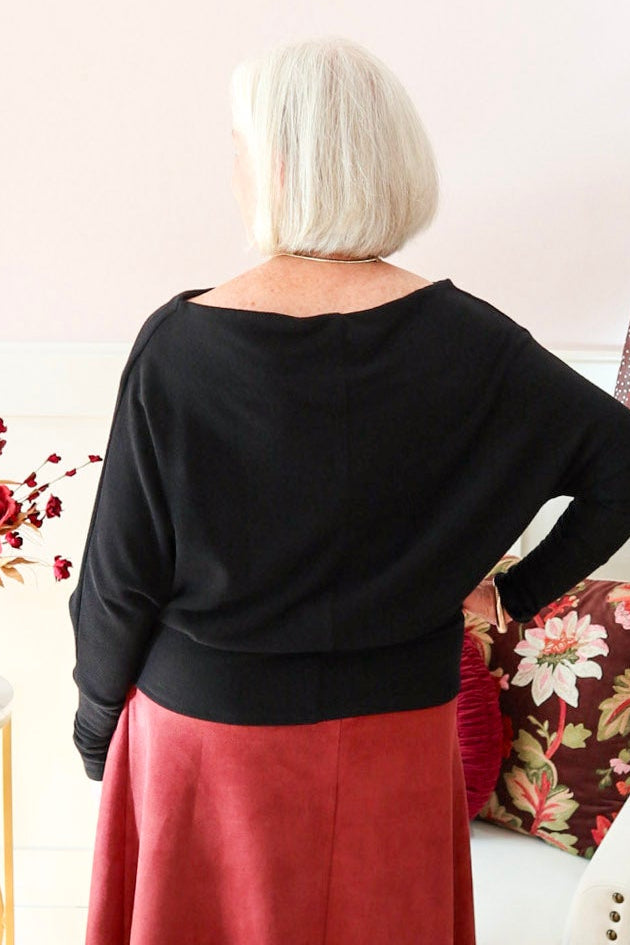 Woman wearing a black Kira knit top with boat neckline and relaxed dolman sleeves, styled with a burgundy suede skirt.