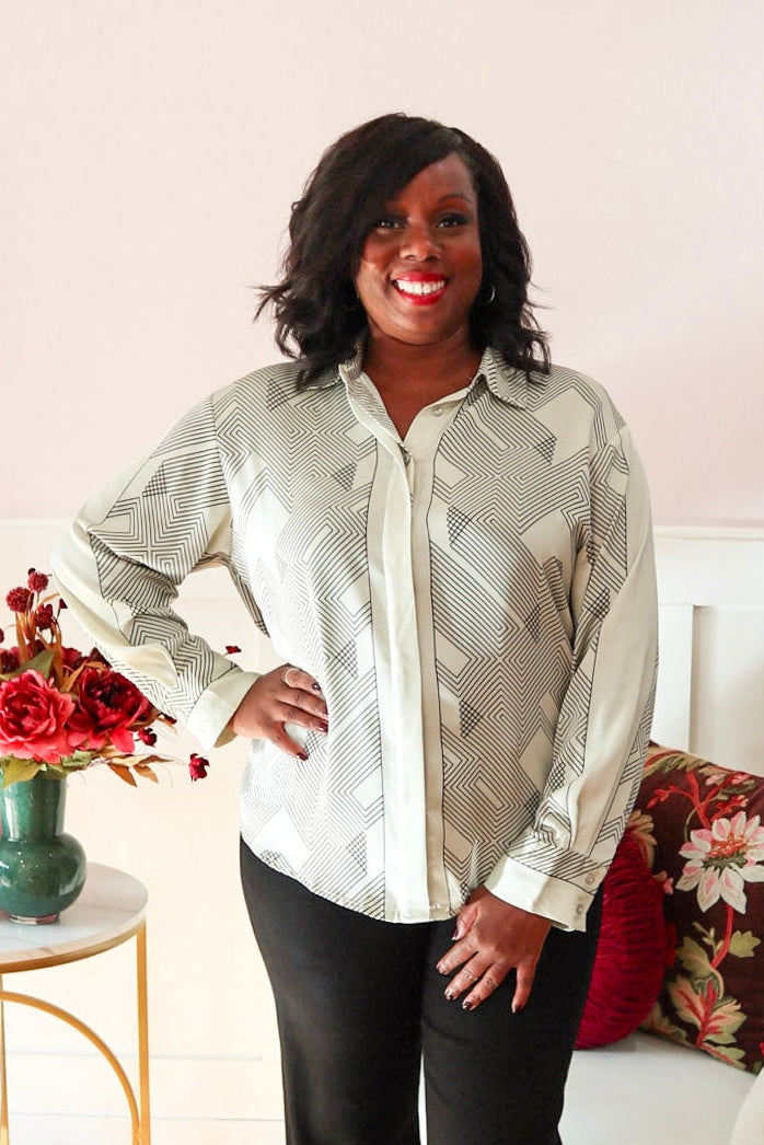 Woman wearing a cream blouse with black geometric print, classic collar, concealed button front, and long sleeves. Styled with black denim jeans. 